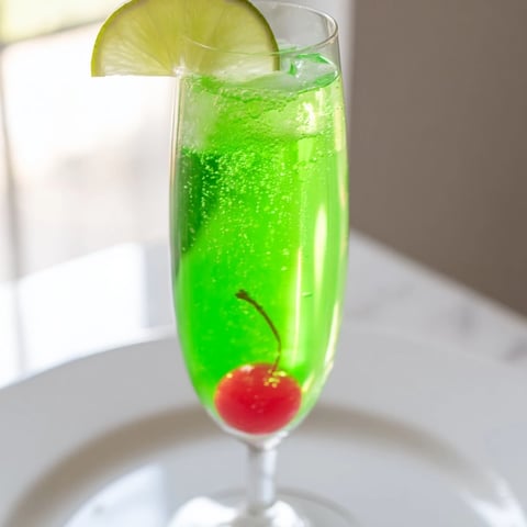 Vibrant green Quick Grinch Mimosa Mocktail in a champagne flute, garnished with cherry and lime.