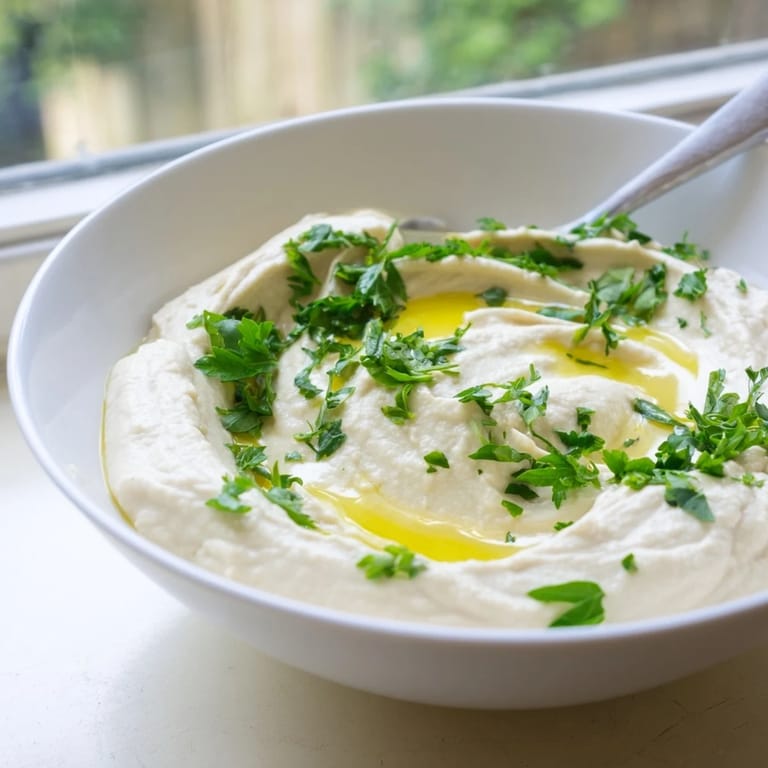 Close-up of smooth White Bean Dip in a white serving bowl, with lemon wedges and fresh garlic cloves nearby on the table.